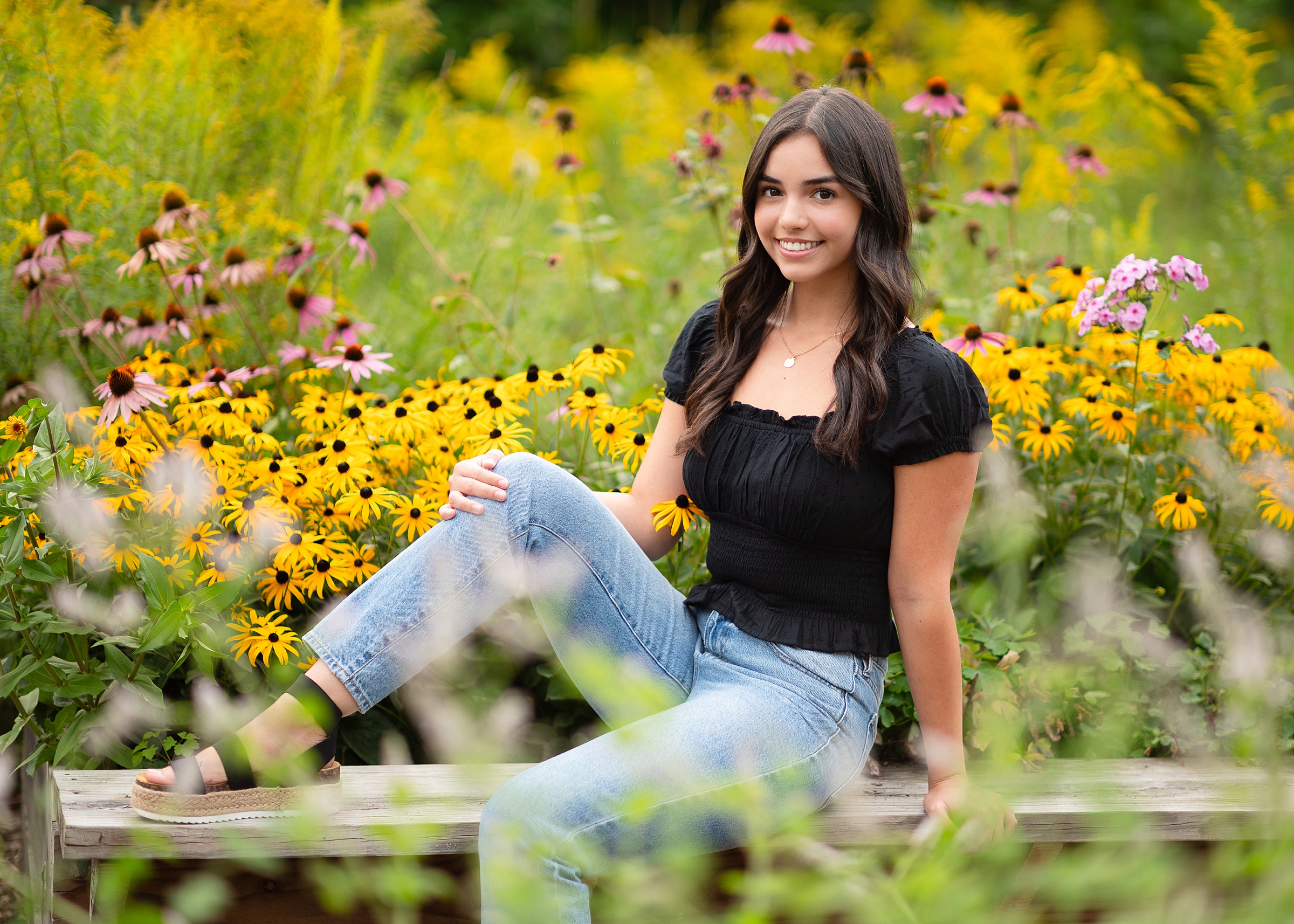 High school senior girl sits on a bench surrounded by wildflowers