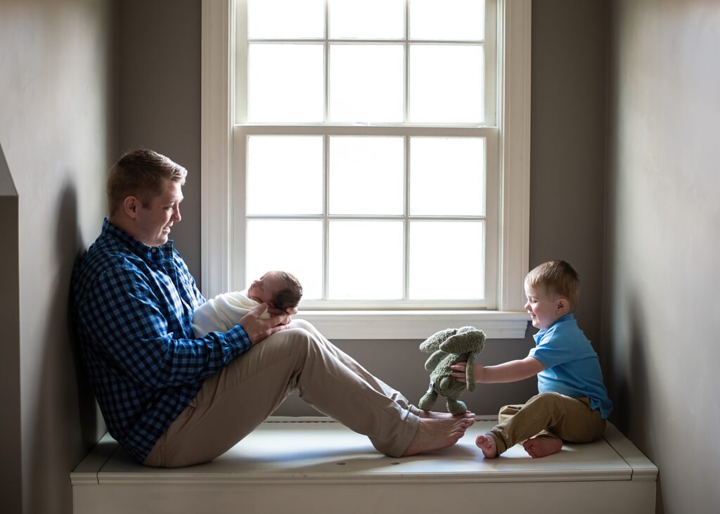 Father holding newborn baby in soft natural light while son plays with stuffed bunny during a Groton MA newborn photography session