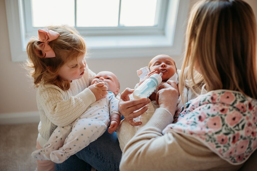 Toddler caring for doll while mom holds baby during in-home newborn photography session